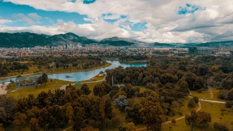 Simón Bolívar Park, a wide aerial view from a drone, showing the treetops, expansive green spaces, the enormous lake, city buildings in the background, and the eastern hills under a cloudy blue sky.
