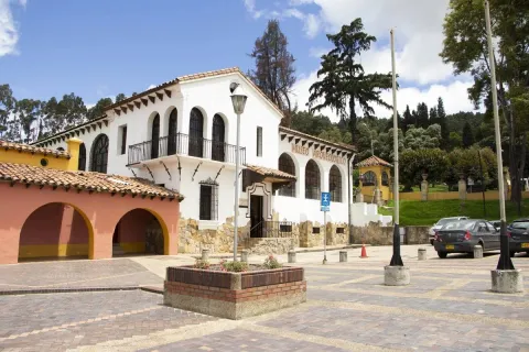 Museo Arqueológico fachada estilo colonial de coor blanco. Al frente una plazoleta con algunos vehículos estacionados y al fondo árboles altos.