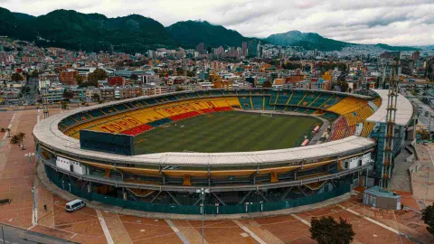 Estadio El Campín en primer plano visto de norte a sur con la ciudad de fondo. El cielo está cubierto de nubes