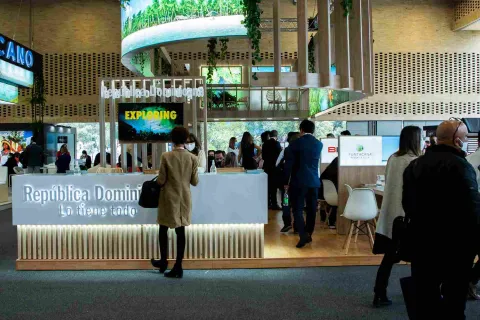  Salón de eventos con un gran stand en el que se lee "República Dominicana". Hay personas alrededor.
