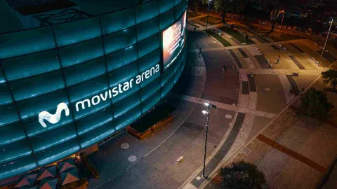 Fachada del movistar Arena de noce con escenario iluminado en azul y con el aviso con letras en neón color blanco. Al frente hay una plazoleta en ladrillo, concreto y vegetación.