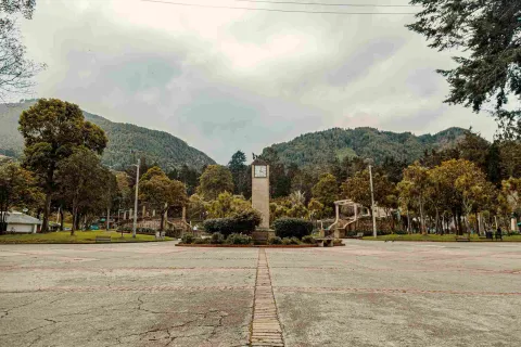 De día una amplia plazoleta con una torre que tiene un reloj en su parte alta. El lugar está rodeado de árboles y al fondo los cerros orientales.