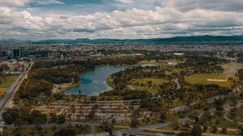 Simón Bolívar Park, a wide aerial view from a drone, looking from east to west, showing green spaces, surrounding trees, the lake in the center, and city buildings in the background. The sky is overcast.