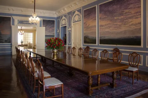 Salon pintado de azul en el que destaca una mesa larga de madera rodeada por sillas, también de madera. En la mesa hay un gran jarrón con flores rojas.
