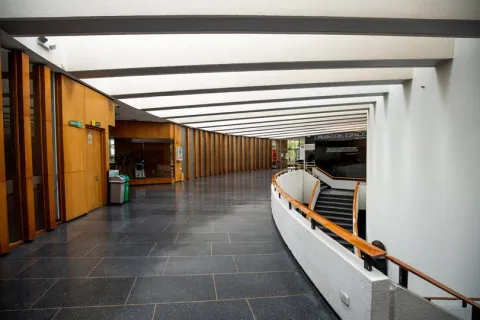  Interior con un salón que parece de biblioteca en el que sobresale un pasillo con paredes en madera, pisos en baldosa negra y paredes blancas. Se ve a la derecha una escalera que conduce a un piso bajo.
