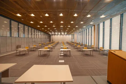 Salón de eventos con grandes ventanales a los lados, luces en el techo, piso entapetado y varias mesas con sillas alrededor.