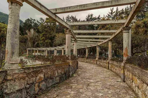 Estructura en piedra tallada y grandes columnas representando una pérgola dividida en dos semicírculos, y rodeada completamente de árboles.