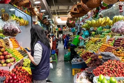 Pasilllo dentro de plaza de mercado con locales atiborrados de frutas. Hay varios vendedores al frente de cada local.