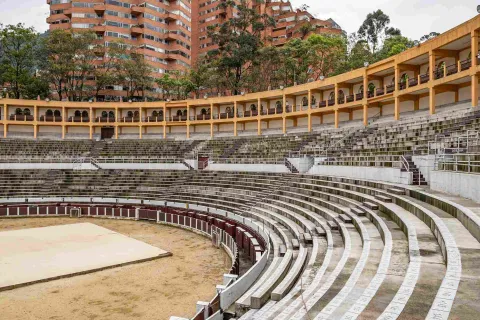Interior de la plaza, se ve un costado de una gradería blanca ovalada de dos pisos y un tercero con balcones de color amarillo. Por encima y al fondo resaltan dos torres de edificios de ladrillo y unos árboles altos.