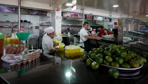 Interior de una amplia cocina en el que un hombre y una mujer adultos están preparando jugos. Se ven varias mandarinas verdes.