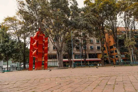 Elcultura metálica alta y de color rojo en una plazoleta en la que al fondo se ve la fachada de varios edificios. 