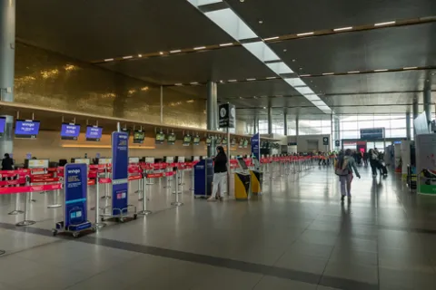 Interior of an airport showing a deep hall with several customer service areas. Some people with suitcases are walking along the front.