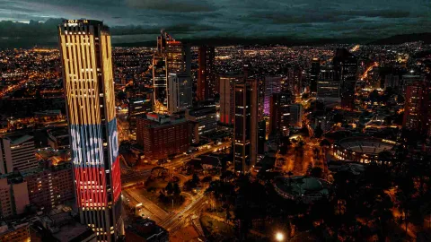 At night, the Colpatria Tower is illuminated with the colors of the Colombian flag. In the background, the lights of the vast city are visible, including several tall buildings and the Santamaría Cultural Center.