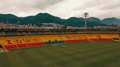 Interior del Estadio, desde arriba, se observa una tribuna de fondo pintada con las palabras IDRD y  Bogotá. Al fondo se alcanzan a ver los cerros orientales. 