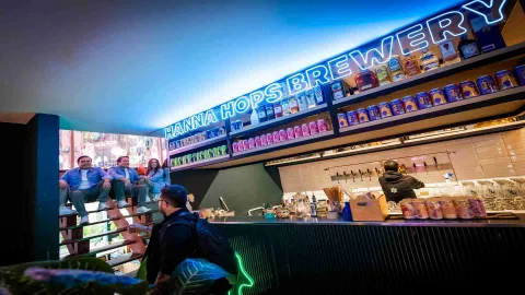 Un salón grande como en una cocina con varios anaqueles con varias botellas y latas de colores, parece que con cerveza. El salón está iluminado por un aviso en neón azul con el nombre del lugar. Hay un hombre y una mujer en una especie de tarima en madera, observando.