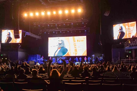 Escenario a oscuras con público sentado y un grupo artístico en la tarima. Se ven tres pantallas gigantes emitiendo un video.