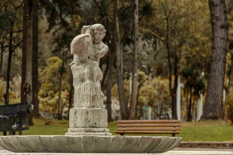 Estructura en piedra tallada rota de un ángel o querubín blanco y que hace parte de una fuente de agua. Al fondo se observan árboles frondosos.