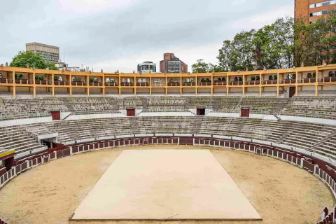 Interior de la plaza, se ve de frente y cerca la arena con una tabla en el centro, al fondo una gradería blanca ovalada de dos pisos; y un tercer piso, con balcones de color amarillo. Por encima y al fondo resaltan la parte alta de algunos edificios y unos árboles altos.