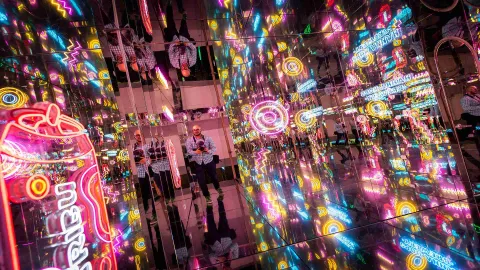 Un hombre joven y calvo con una cámara fotográfica en medio de un pasillo con paredes de espejos de cristal iluminados con luces de neón de varios colores. 