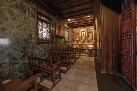 Interior de capilla con paredes y techos de madera y mármol. Hay una ventana en vitral cinco sillas de madera amplias y vacías. Al fondo hay un altar.