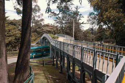  Puente de hierro con figuras geométricas sobre una avenida por la que pasa rápido un bus. Está rodeado de altos árboles.