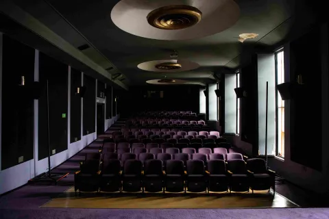 Salón como de cine con varias filas de sillas color violeta, de frente a fondo, lámparas inclrustadas en el techo y a un lado 4 ventanas abiertas con luz de día afuera.