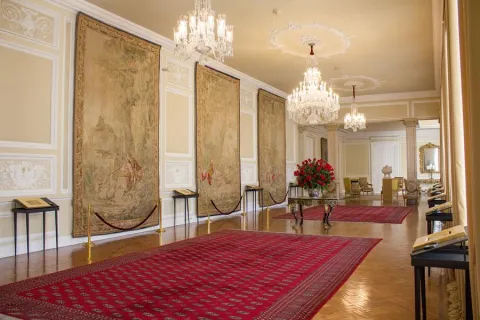Salón largo con tapetes rojos y 3 tapices en una pared. Hay tres lámparas de cristal en el techo.