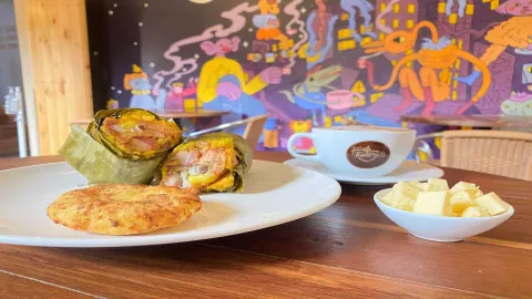 Un tamal envuelto en hoja, una arepa de maíz, una taza de café y un tazón de queso, en una mesa de madera.