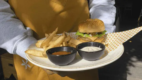 Hamburguesa con papas fritas, kétchup y mayonesa en recipientes negros, servida en plato blanco.