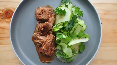 Un plato con carne desmenuzada en salsa y una ensalada fresca de pepino y hojas verdes.