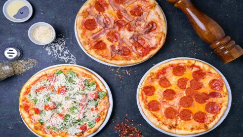 Tres pizzas deliciosas: una de pepperoni, otra de jamón, y una vegetariana, en una superficie oscura con especias.