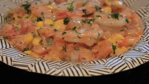 Ceviche cremoso con camarones, trozos de elote y tomate, salpicado con perejil, en un plato estampado.