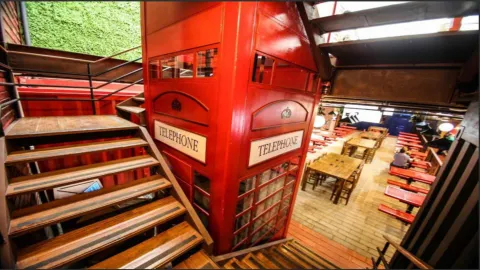  Interior de un restaurante con una cabina telefónica roja icónica, escaleras de madera y mesas de madera.