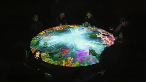  Comensales alrededor de una mesa redonda con proyecciones interactivas de vida marina, en un ambiente oscuro.