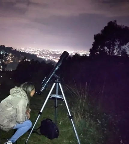 Mujer joven con una chaqueta clara observa de noche a través de un telescopio dirigido hacia las estrellas.