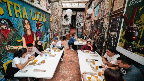 Grupo de persons jóvenes en un corredor con las paredes llenas de grafitis, sentados al rededor de mesas alargadas y consumiendo platos típicos con arroz, patacones y jugo. 