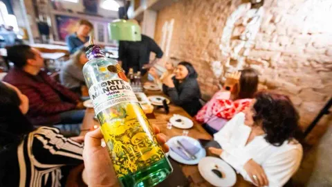 Primer plano de botella de licor en la que la etiqueta dice que es viche. Al fondo borroso se ve a un grupo de personas sentados alrededor de una mesa.