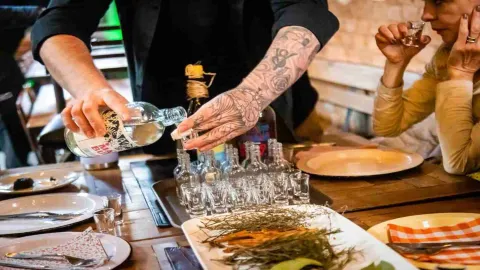Una persona con manos tatuadas sirve en una copa el licor de una botella. Está al frente de una mesa en la que hay varias copas pequeñas y algunos vegetales.