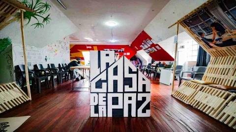 Un salón con un aviso de madera grande en el centro con la figura de una casa y el título en letras blancas y mayúsculas "LA CASA DE LA PAZ". El salón está rodeado por sillas y decoraciones de madera en las paredes.