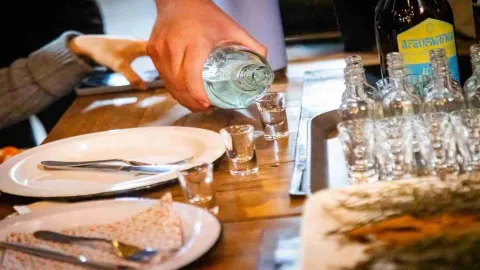 En primer plano una mano sirve licor de una botella en unas copas pequeñas.