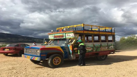  Bus pintado de colores denominado como la tradicional chiva. Tiene ventanas sin vidrios y un segundo piso sin techo. Un hombre, que parece el conductor intenta subir.