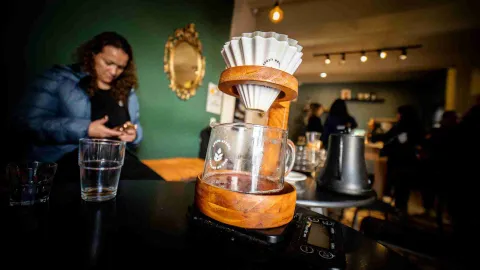 Primer plano de artefacto de madera para hacer café, con un filtro en papel blanco. Al fondo se oberva a una mujer observando un teléfono móvil.