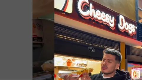 Primer plano de un hombre joven comiendo un trozo de perro caliente. Al fondo se observa un aviso con una leyenda que dice: "Cheesy Dogs".