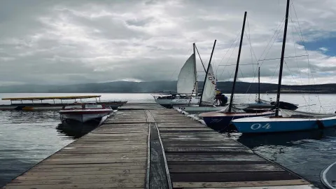 Muelle en un lago con varios veleros alrededor. El cielo está nublado.