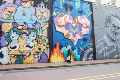 Calle en la que se observan tres paredes pintadas con graffitis. En una hay un elefante psicodélico; en otra corazones de colores; y en otra en blanco y negro un rostro de un hombre anciano. Una mujer y una niña caminan por el frente.