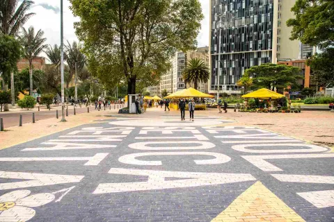 Calle con avisos pintados en el piso y por la que van pasando algunas personas. Se observa al fondo algunos toldos amarillos, y una caseta con un guarda de seguridad.