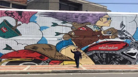  Hombre joven de pie al frente de un grafiti en el que se ve a una mujer joven que va en una motocicleta.