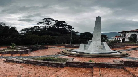 Plaza enladrillada con un obelisco no muy alto  de piedra color blanco. Al fondo se ven varios árboles frondosos.