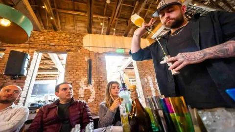 Dos hombres y una mujer jóvenes atiende a otro hombre que parece ser un barman, y que hace una pirueta con el líquido de una botella a una copa de metal.
