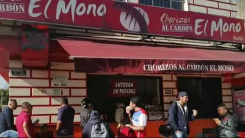 Personas al frente de un local comercial comiendo. Hay un aviso con una leyenda que dice: "Chorizos el Mono".